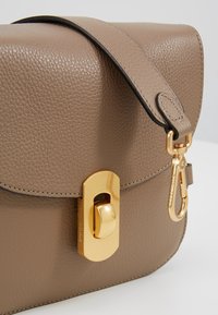 Bolso bandolera de cuero beige con superficie texturizada, herrajes dorados, un cierre prominente y un borde negro contrastante en la correa.