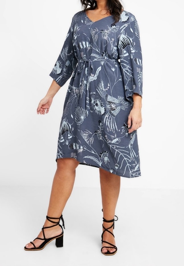 Femme portant une robe bleue mi-longue avec des motifs floraux et d'oiseaux blancs, manches trois-quarts, lien à la taille et sandales noires à talons blocs et à brides.