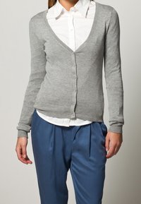 Grau, langärmlige Strickjacke mit V-Ausschnitt, Knopfleiste und schmalem Bund. Kombiniert mit blauen gefältelten Hosen und einem weißen Hemd mit Kragen.