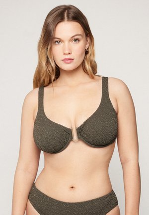 Junge Frau mit langen blonden Haaren, die einen strukturierten dunkelolivgrünen Bikini mit goldener Frontklammer trägt und vor einem einfarbigen hellen Hintergrund posiert.