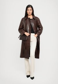 Donna che indossa un trench lungo in pelle marrone scuro, top nero, pantaloni color crema, tacchi neri e porta una borsa a spalla marrone scuro coordinata.