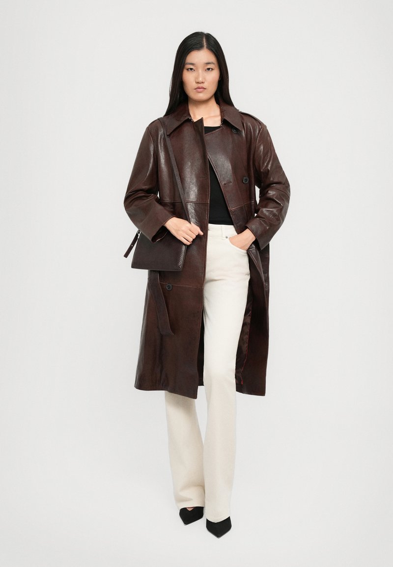 Donna che indossa un trench lungo in pelle marrone scuro, top nero, pantaloni color crema, tacchi neri e porta una borsa a spalla marrone scuro coordinata.