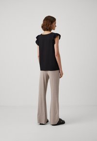 Svart topp med volangermet ermer, sammen med lyse beige, teksturerte vide bukser. Modellen står foran en ensfarget grå bakgrunn.