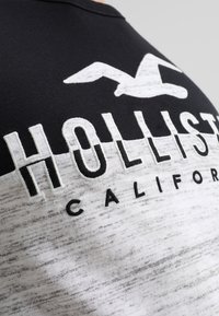 Musta ja valkoinen lyhythihainen T-paita, jossa on graafinen printti "HOLLISTER CALIFORNIA" ja lokin logo rinnan alueella.
