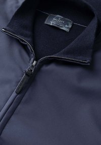 Tmavomodrá bunda s polovičným zipsom, rebrovaným golierom a štítkom Hackett London vo vnútri goliera s nápisom „Tailored for Travel“ (Šitá na mieru pre cestovanie).