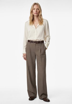 Woman standing wearing a cream long-sleeve blouse tucked into high-waist brown wide-leg trousers with a dark belt and brown shoes.