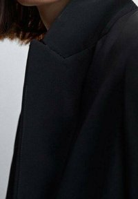 Una giacca nera con un colletto strutturato, caratterizzata da una superficie liscia e linee pulite. Il tessuto sembra leggero e su misura.