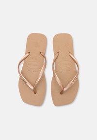 Tongs en caoutchouc tan avec des sangles en métal rose doré, semelle intérieure texturée et détail de logo sur la sangle, présentant un design minimaliste.