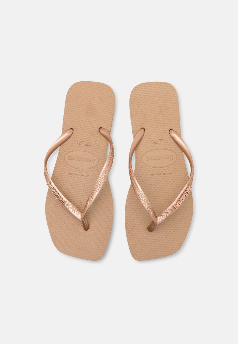 Tongs en caoutchouc tan avec des sangles en métal rose doré, semelle intérieure texturée et détail de logo sur la sangle, présentant un design minimaliste.
