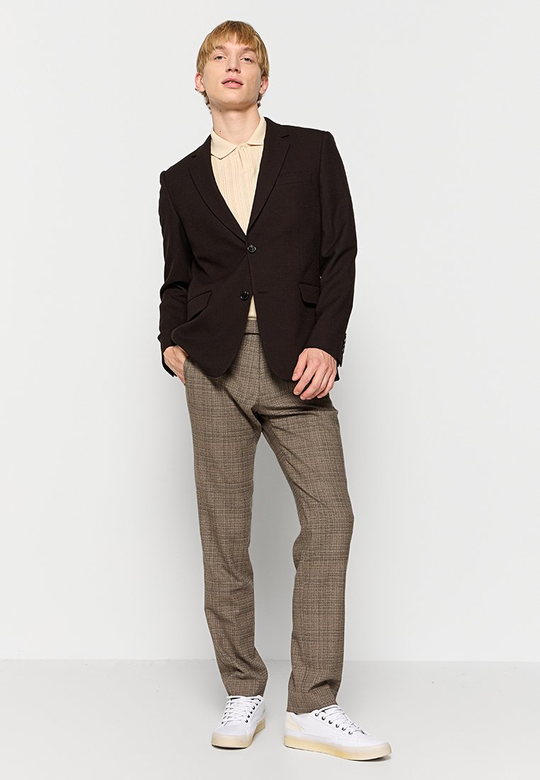 Brauner maßgeschneiderter Blazer mit zwei Knöpfen, kombiniert mit einem beigefarbenen Hemd mit Kragen und braunen karierten Hosen. Weiße Sneakers vervollständigen das Outfit.