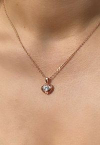 Collana con pendente a forma di cuore in oro rosa, dotata di una gemma centrale trasparente, appesa a una catena delicata, che poggia sulla pelle.
