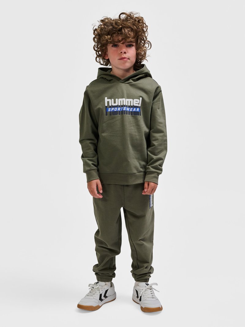 Dziecko o kręconych włosach, ubrane w oliwkową bluzę z kapturem "hummel SPORTSWEAR", dopasowane spodnie i białe trampki z czarnymi chevronami, stoi nieruchomo.