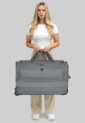 Giovane donna con lunghi capelli biondi tiene una grande borsa a rotelle grigia con due manici contro uno sfondo grigio chiaro.