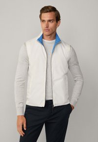 Vit ärmlös väst med blå inre krage, bärs över en ljusgrå långärmad skjorta, tillsammans med mörka byxor. Slätt, lätt material.