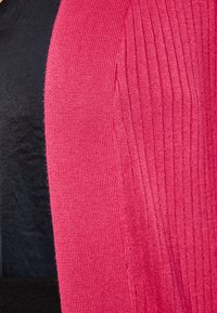 Růžová hřejivá pletenina vpravo kontrastuje s hladkým černým materiálem vlevo, což ukazuje texturální a barevné rozdíly.