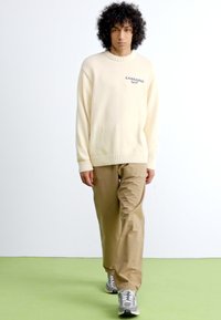 Jeune personne avec des cheveux bouclés marchant, portant un pull crème, un pantalon beige et des baskets grises sur un sol vert clair.