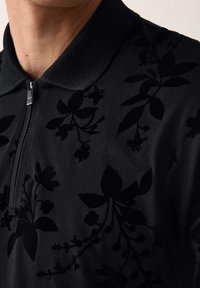 Közelkép egy fekete cipzáros ingről, amelyen texturált virágminta és bordázott gallér látható egy személy mellkasának és nyakának felső részén.