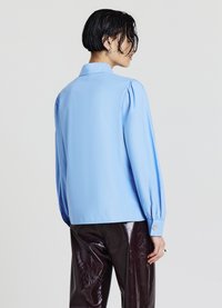 Light blue blouse with a collared neckline and puffed long sleeves. Smooth fabric with a slightly shiny finish, worn with dark, glossy pants.