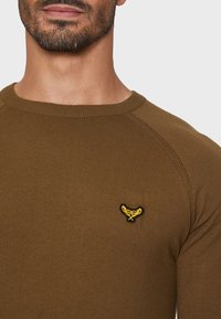 Brun ribbet sweater med rund halsudskæring og sort-gul broderet logo på brystet. Har raglanærmer. Blød tekstur.