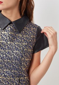 Vestido de fondo marino y dorado con un cuello de cuero negro y mangas cortas de cuero. Presenta un diseño texturizado y adornado.