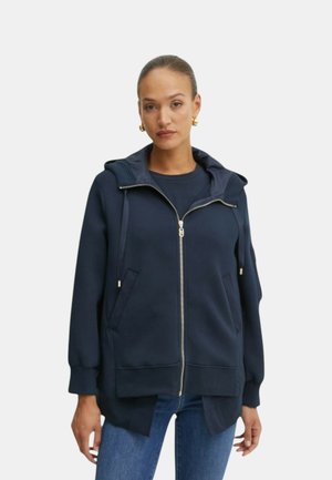 Felpa con zip blu navy, dotata di cappuccio, tasche frontali e dettagli in metallo dorato. Il tessuto morbido presenta una superficie texturizzata e una vestibilità comoda.