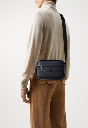 Persona que lleva un jersey de cuello alto beige y pantalones marrones, y lleva un bolso bandolera negro de Karl Lagerfeld con logo en relieve y correa ajustable.