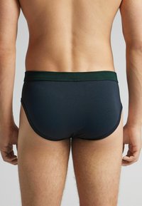 Bakifrån av en person som bär mörkblå kalsonger med ett grönt midjeband mot en enfärgad bakgrund.