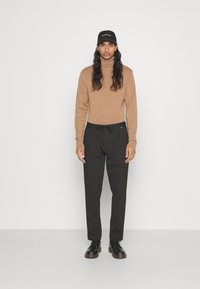 Felpa dolcevita marrone chiaro, pantaloni neri con cordino e scarpe nere; modello con capelli lunghi che indossa un cappellino nero, in piedi contro uno sfondo bianco.