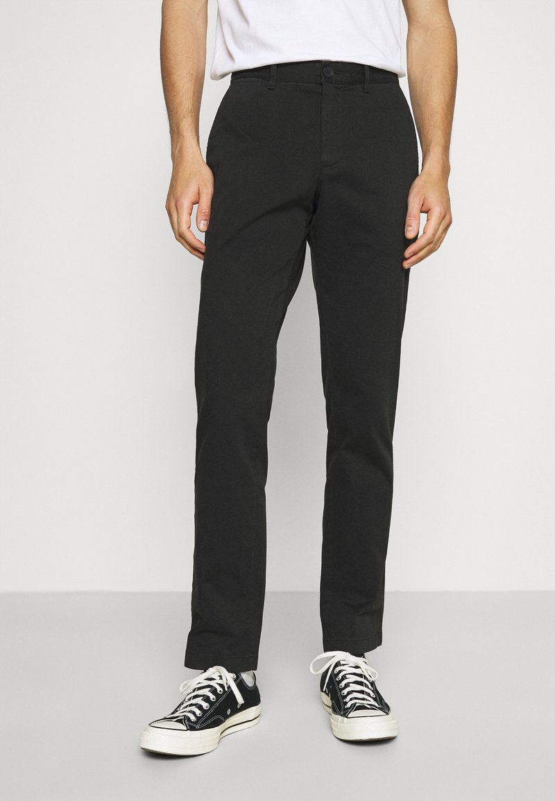 Homme portant un pantalon noir droit, des baskets noires et blanches et une chemise blanche, debout devant un fond clair uni.
