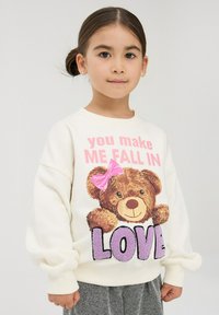 Kremowy sweter z grafiką misia z różową kokardą oraz napisem "sprawiasz, że zakochuję się" w kolorze fioletowym i czarnym. Miękka tkanina, luźny krój.