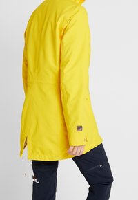 Osoba w jasnożółtej kurtce przeciwdeszczowej z regulowanymi sznurkami i czarnych spodniach z zamkiem, pokazana od tyłu na neutralnym tle.