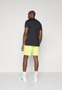 T-shirt de sport noir à manches courtes avec un logo sur la manche gauche, associé à un short jaune fluo. Baskets blanches aux pieds.