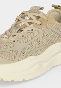Basket de sport beige et crème avec tige en mesh et synthétique, semelle blanche épaisse, lacets beiges et détails de marque sur la languette et la zone des orteils.