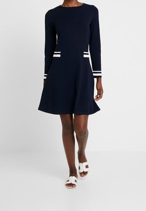 Kvinde går i en marineblå, langærmet kjole med hvide stribede accenter og hvide sandaler med åben tå mod en ensfarvet baggrund.