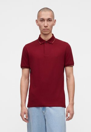 Bărbat cu capul ras, purtând un tricou polo roșu închis și blugi albastru deschis, stând în picioare în fața unui fundal alb simplu.