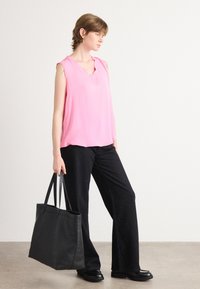Mouwloze roze blouse met een V-hals, gecombineerd met zwarte, glinsterende wijde broekspijpen. Model houdt een zwarte tote bag vast. Soepele stofstructuur.