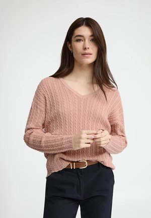 Młoda kobieta o długich brązowych włosach, ubrana w różowy sweter w splot przerzutowy z dekoltem w serek oraz czarne spodnie z brązowym paskiem, stojąca na tle białej ściany.