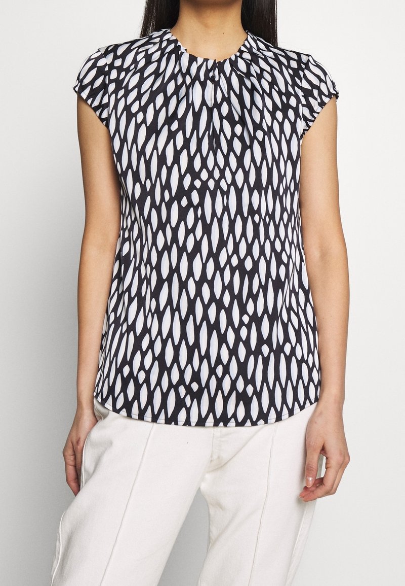 Kvinna klädd i en ärmlös blus med svart och vit mönstring med plisseringar vid kragen, i kombination med off-white byxor, stående mot en enfärgad bakgrund.