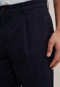 Temno modri hlače iz lahkega materiala, s ploskim sprednjim delom, stranskimi žepi in gladko teksturo. Gumbasto zapiranje v pasu.