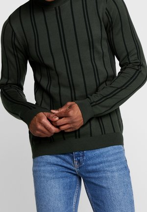 Mann trägt dunkelgrünen Pullover mit vertikalen schwarzen Streifen und blaue Jeans, Hände vor der Taille verschränkt.