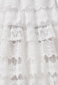 Tissu en dentelle blanche avec motifs floraux et géométriques superposés sur matériau transparent, montrant une broderie délicate et des bords festonnés.