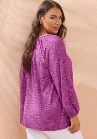 Lila, übergroße Bluse mit einem strukturierten, dezenten Zebra-Muster, ballonförmigen Ärmeln und einem runden Ausschnitt, kombiniert mit weißen Hosen.