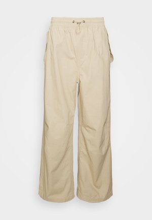 Person iført lys grå lynlås-hoodie, hvid grafisk t-shirt, beige bukser og hvide sneakers, stående på en træstol.