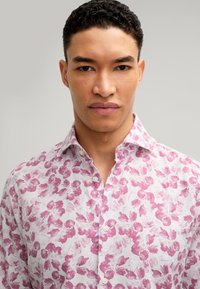 Weißes Hemd mit pinkem Blumenmuster, Button-down-Kragen und weicher Textur. Nahaufnahme des Oberkörpers vor neutralem Hintergrund.