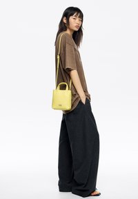Żółta skórzana torebka z krótkimi i długimi rączkami, zestawiona z oversize'ową brązową koszulką i szerokimi szarymi spodniami. Prosty design, minimalistyczne akcenty.