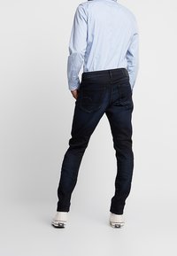 Mörka denimjeans med slim fit, med bakfickor och midjeband. Bärs med en ljusblå skjorta och vita sneakers.