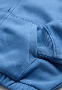 Blauwe sweatshstoff-detail met geribde manchetten, kangoeroezak en zichtbare stiksels op zacht, gestructureerd materiaal.