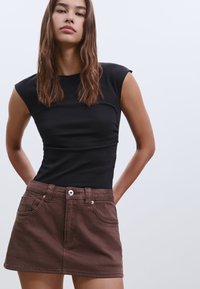 Schwarzes, figurbetontes, ärmelloses Oberteil kombiniert mit einem braunen Denim-Minirock, der über frontale Taschen und einen silbernen Knopfverschluss verfügt.