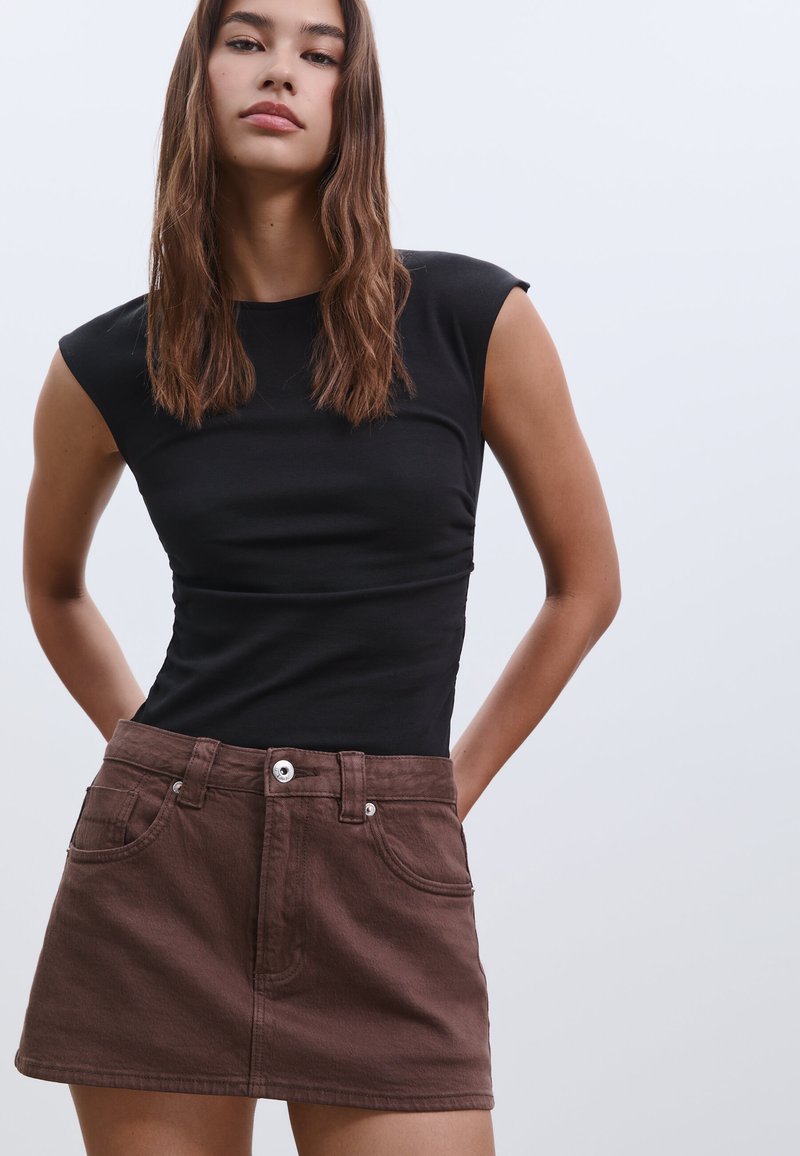 Schwarzes, figurbetontes, ärmelloses Oberteil kombiniert mit einem braunen Denim-Minirock, der über frontale Taschen und einen silbernen Knopfverschluss verfügt.