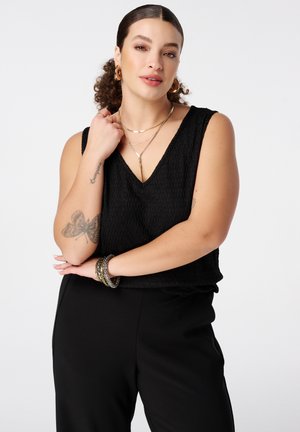 Vrouw met krullend haar, zwarte mouwloze top en broek, draagt gouden sieraden en heeft een vlindertattoo op de linker onderarm.